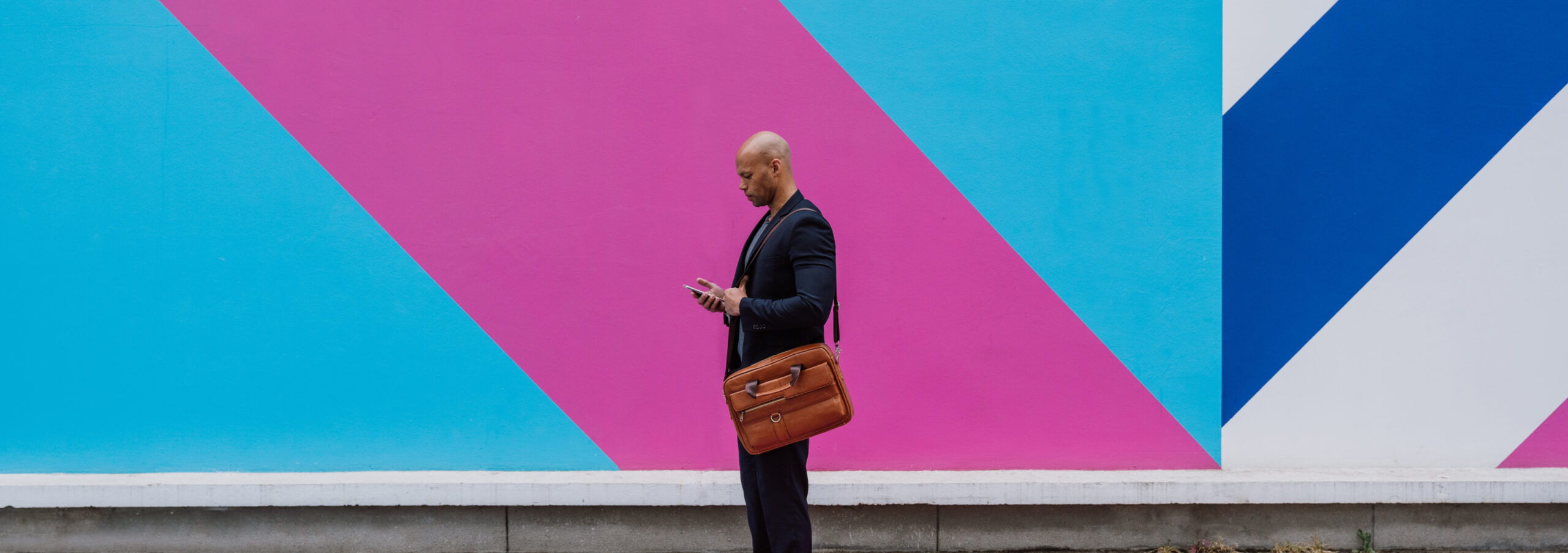 Man in a business suit with an over the shoulder bag standing on sidewalk looking at their phone